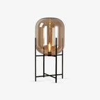 Amber glass shade with a visible filament bulb on a slim black four-leg stand, this Clérachic lamp on a plain light grey background.