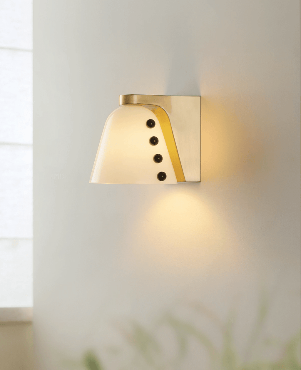 The white glass shade glows warm on a pale wall, casting a soft pool of light downward, with blurred grass in the foreground; a Clérachic wall light.