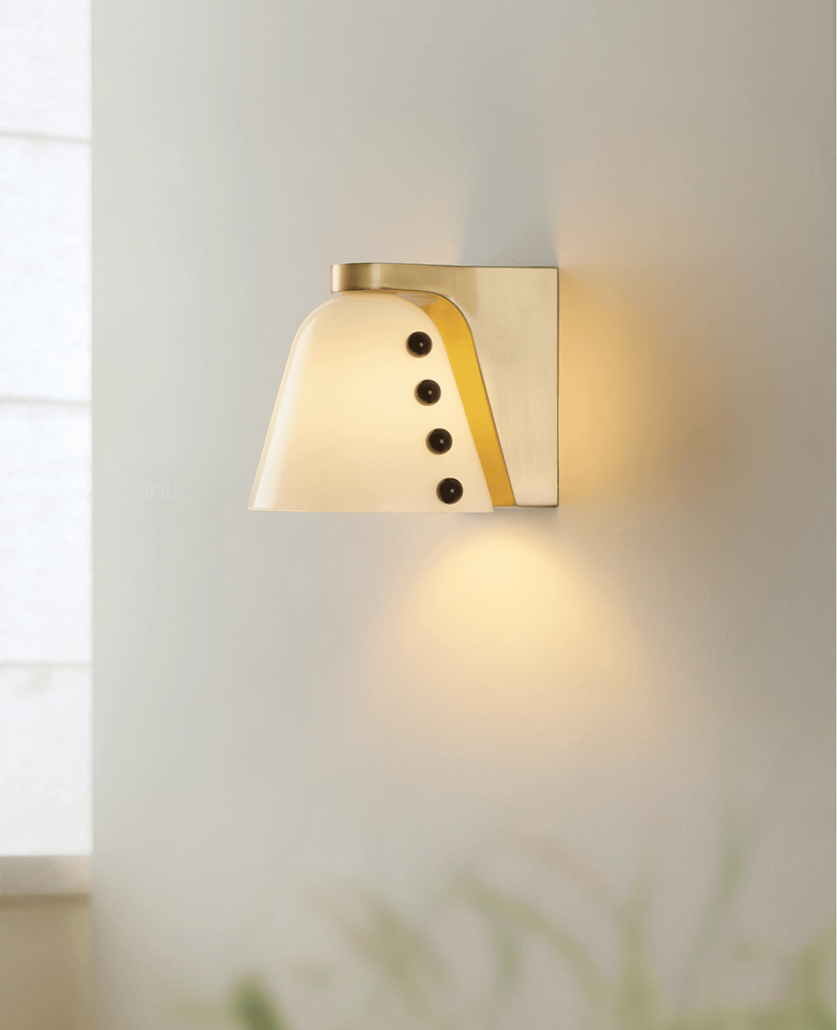 The white glass shade glows warm on a pale wall, casting a soft pool of light downward, with blurred grass in the foreground; a Clérachic wall light.