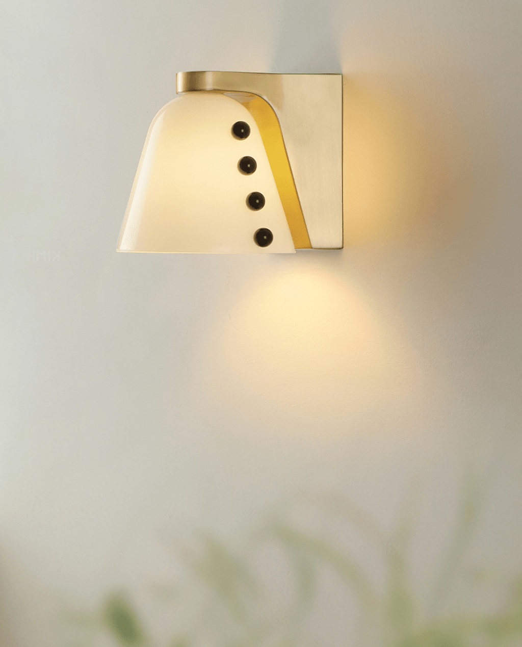 Close, lit view of the white glass shade with a gold backplate and four black studs, light spilling below; a Clérachic wall light.