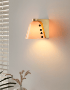 A shade in a peach-orange glass lit against a grey wall near window blinds and dried pampas stems; a Clérachic wall sconce.