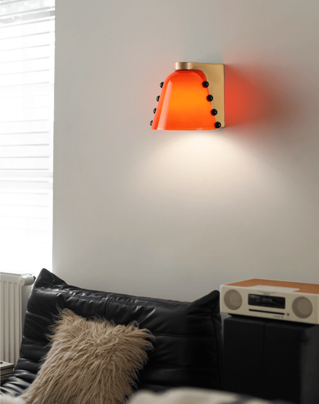 Deep red-orange glass shade lit above a black leather sofa, a fluffy cushion and a retro radio; a Clérachic wall light.