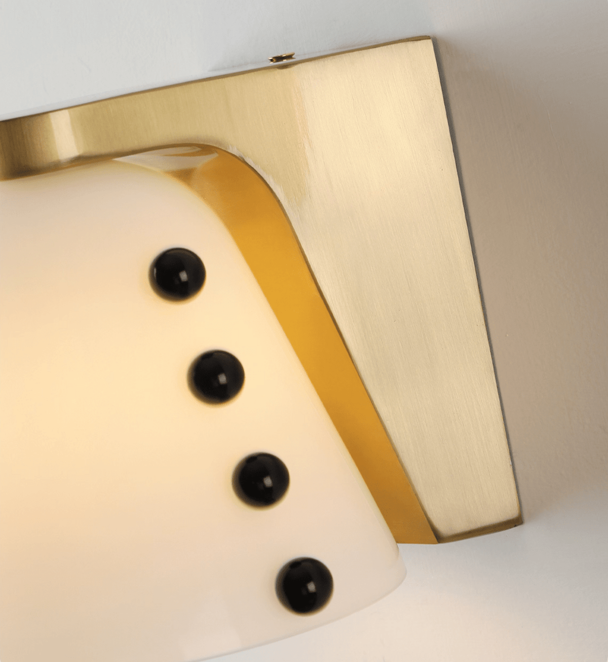 Very close view of the brushed gold bracket, the white glass and four glossy black studs on this Clérachic wall light.