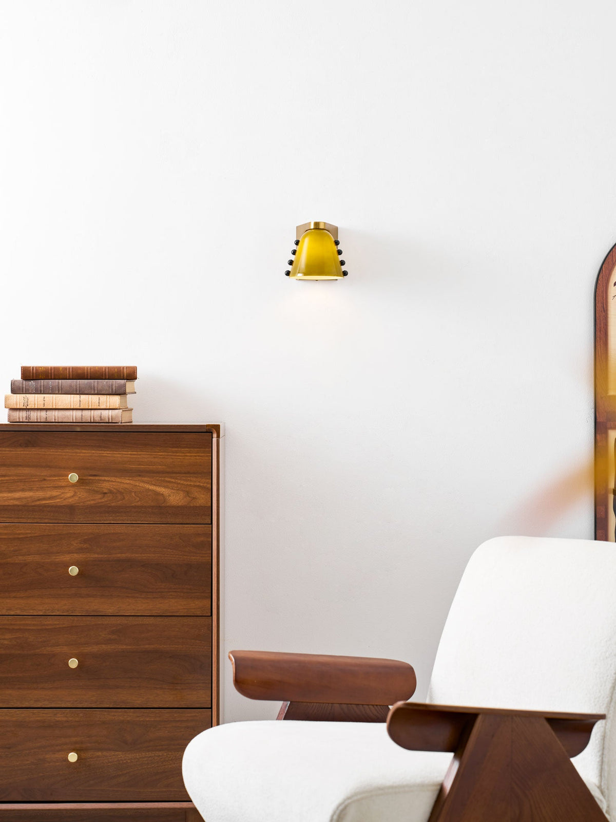 A small lit wall light above a walnut chest of drawers topped with books, beside a white armchair; from Clérachic.