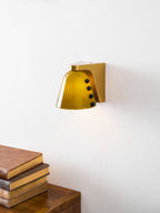 Green glass wall light with black studs and gold bracket above a stack of books and a walnut cabinet; a Clérachic design.
