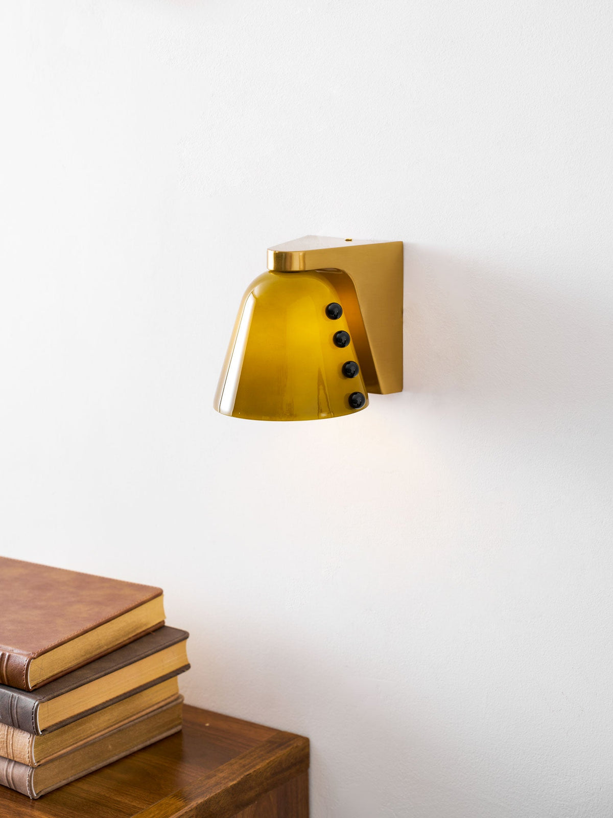 Green glass wall light with black studs and gold bracket above a stack of books and a walnut cabinet; a Clérachic design.