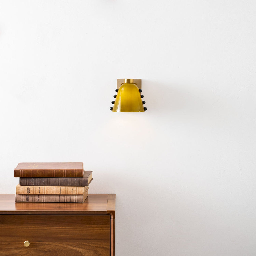 Wider view of the green glass sconce with a soft glow, over a book stack on a wooden chest; from Clérachic.