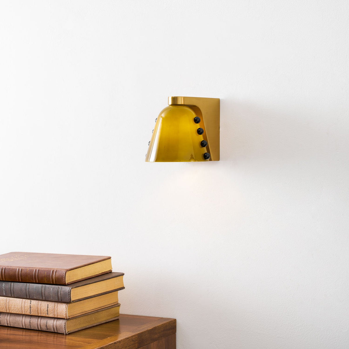 Green glass shade lit against a white wall, with books and a wooden surface in the lower corner; a Clérachic wall sconce.