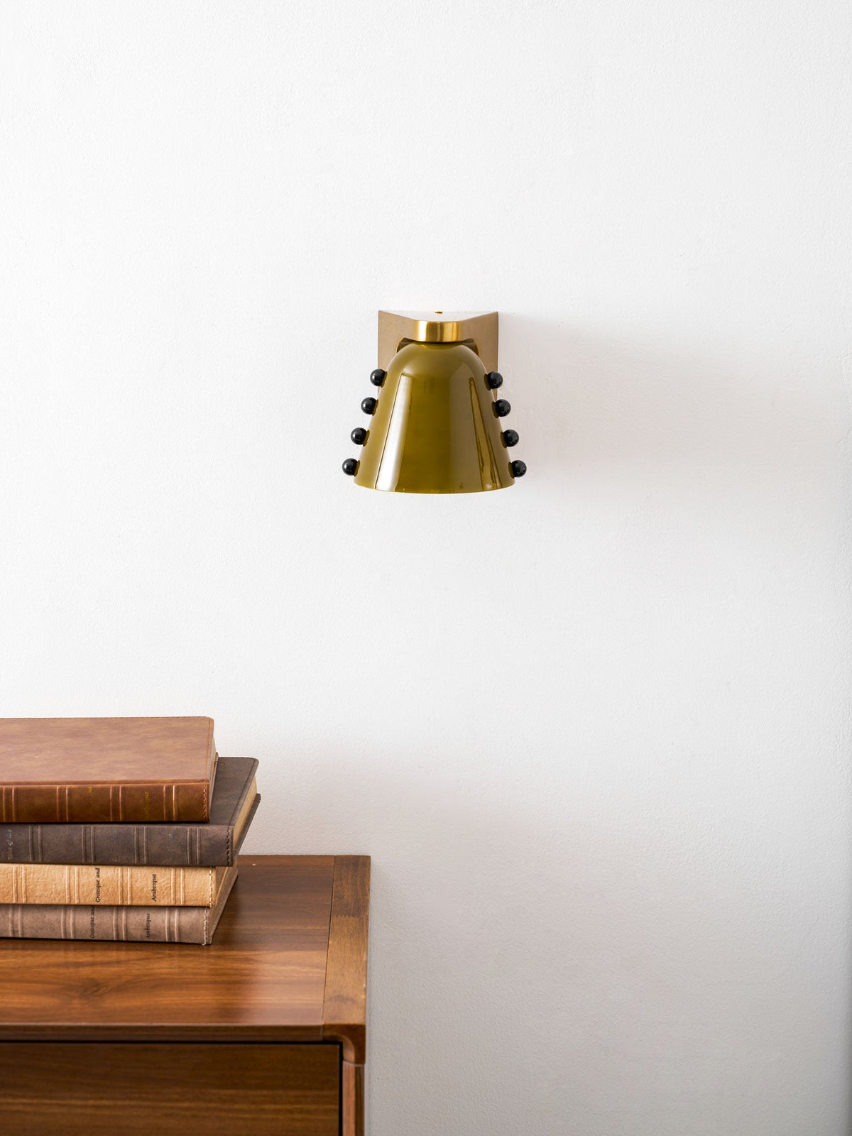 Green glass wall light seen with its studs on both sides, above a wooden cabinet and a pile of books; from Clérachic.