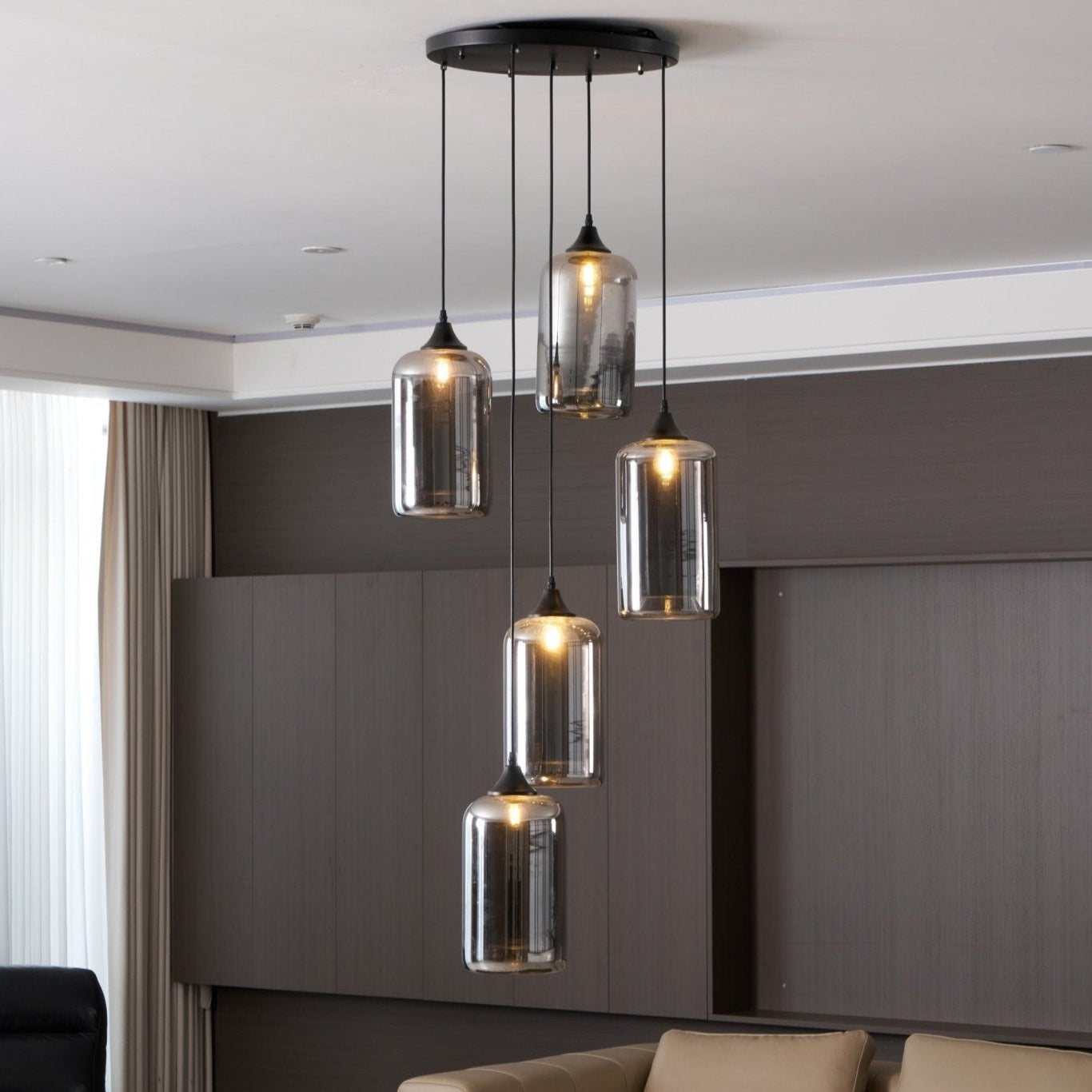 Five cylinder Clérachic smoked glass pendants from a round canopy, lit above a panelled wall and sofa.