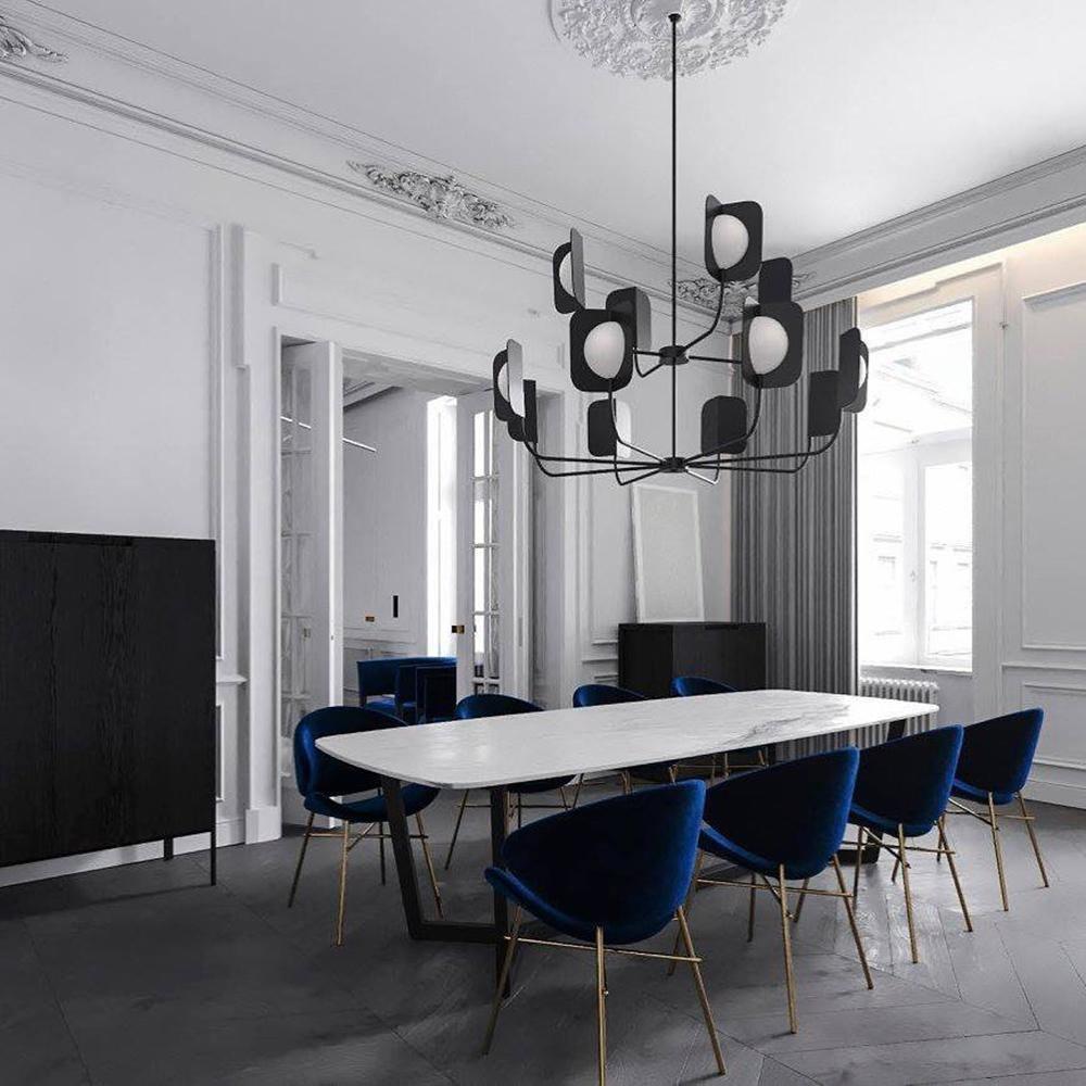 A black Clérachic chandelier hangs over a marble dining table with deep blue velvet chairs, in a white room with a carved ceiling rose.