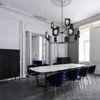 A black Clérachic chandelier hangs over a marble dining table with deep blue velvet chairs, in a white room with a carved ceiling rose.