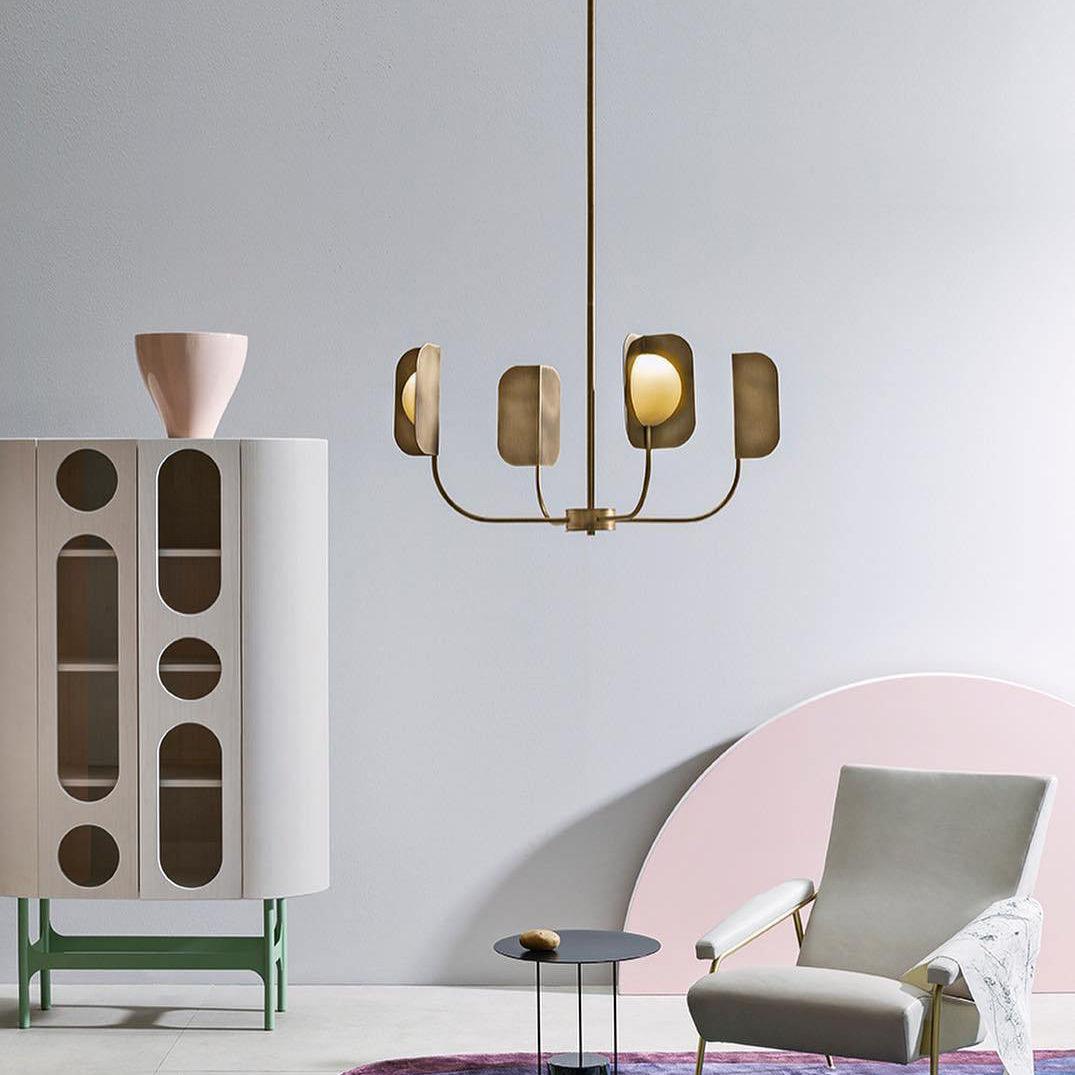 A four-arm Clérachic chandelier in a brass-toned finish hangs beside a tall cabinet with arched cut-outs and a pale grey armchair.