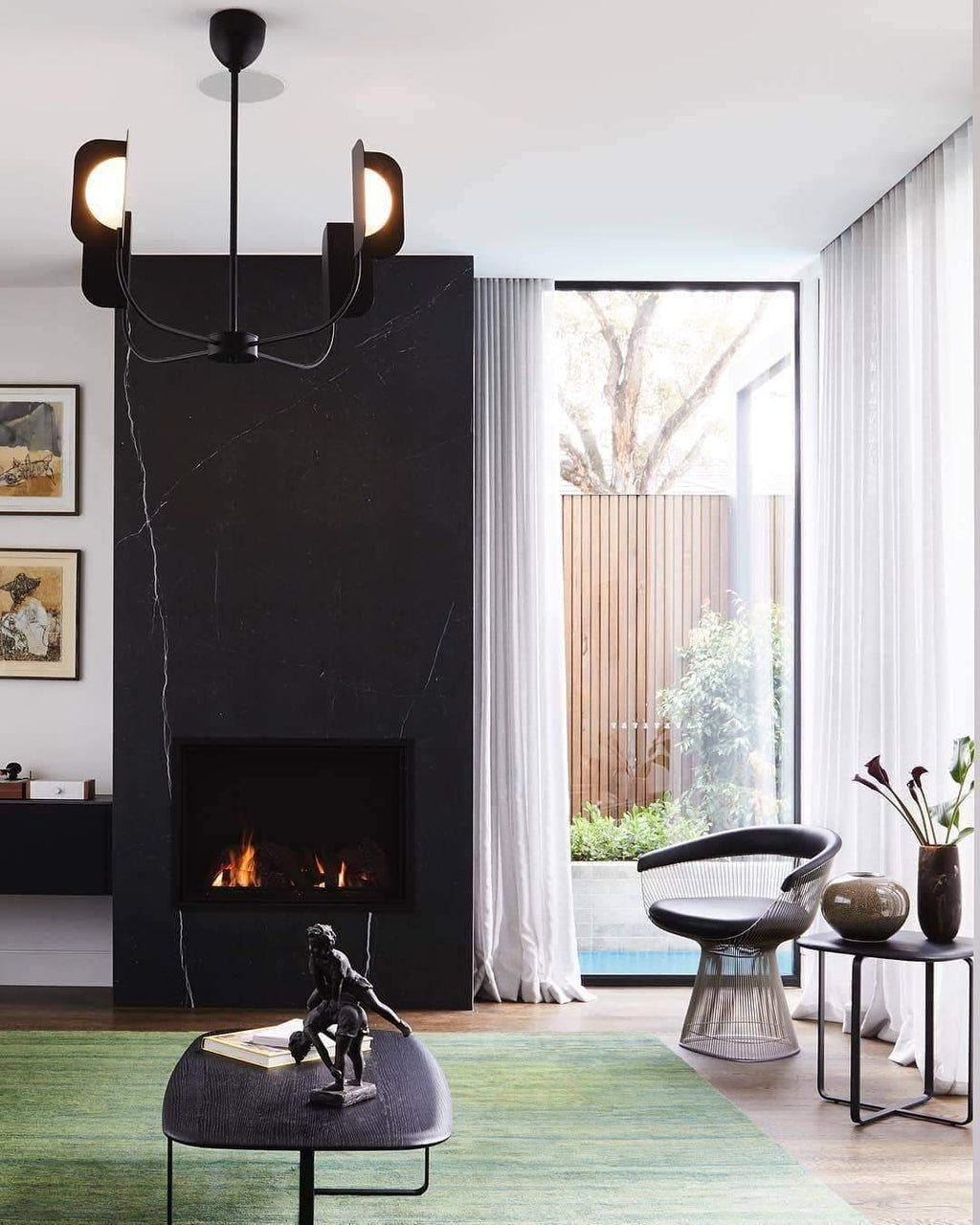 A black four-arm Clérachic chandelier hangs in front of a black stone fireplace, with sheer curtains and a garden beyond the glass.