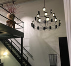 Lit black Clérachic chandelier in a double-height room with a black steel stair, its tiers of leaf shades glowing warm from the globes.