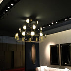 A brass-toned Clérachic chandelier lit in a dark showroom, three tiers of arms with leaf shades and globes beside a gold-framed mirror.