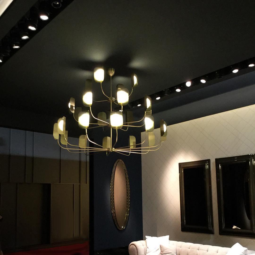 A brass-toned Clérachic chandelier lit in a dark showroom, three tiers of arms with leaf shades and globes beside a gold-framed mirror.