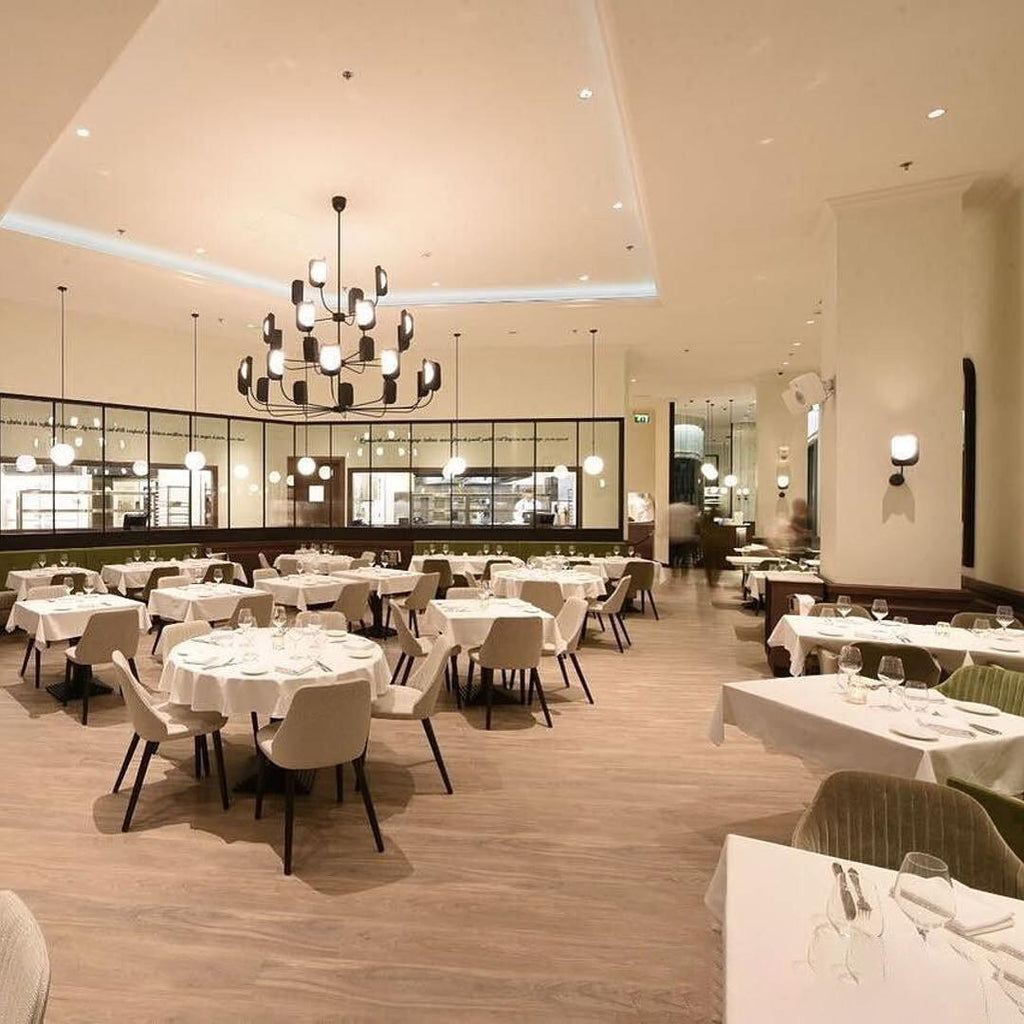 A black Clérachic chandelier lit high in a restaurant dining room with white tablecloths, its leaf shades glowing above the tables.