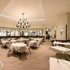 A black Clérachic chandelier lit high in a restaurant dining room with white tablecloths, its leaf shades glowing above the tables.