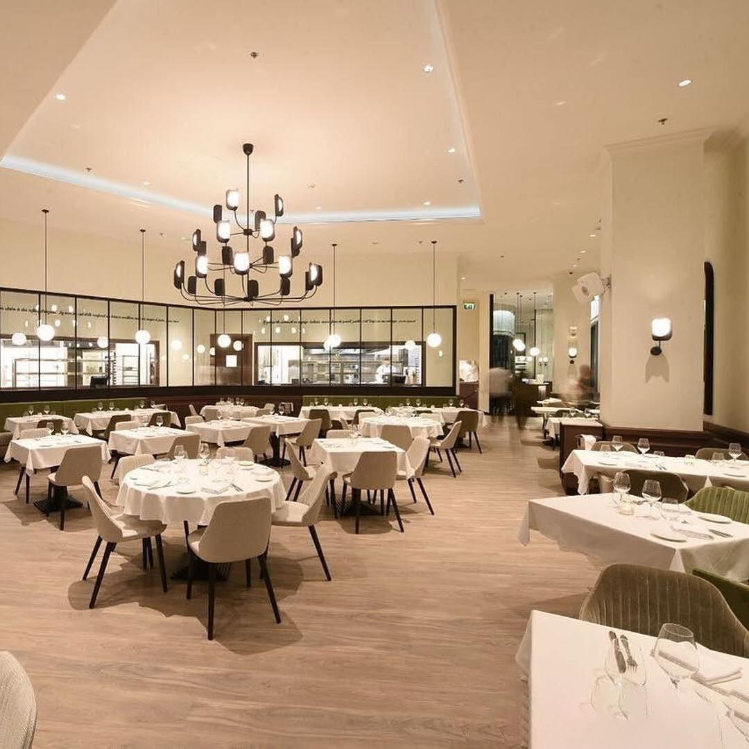 A black Clérachic chandelier lit high in a restaurant dining room with white tablecloths, its leaf shades glowing above the tables.