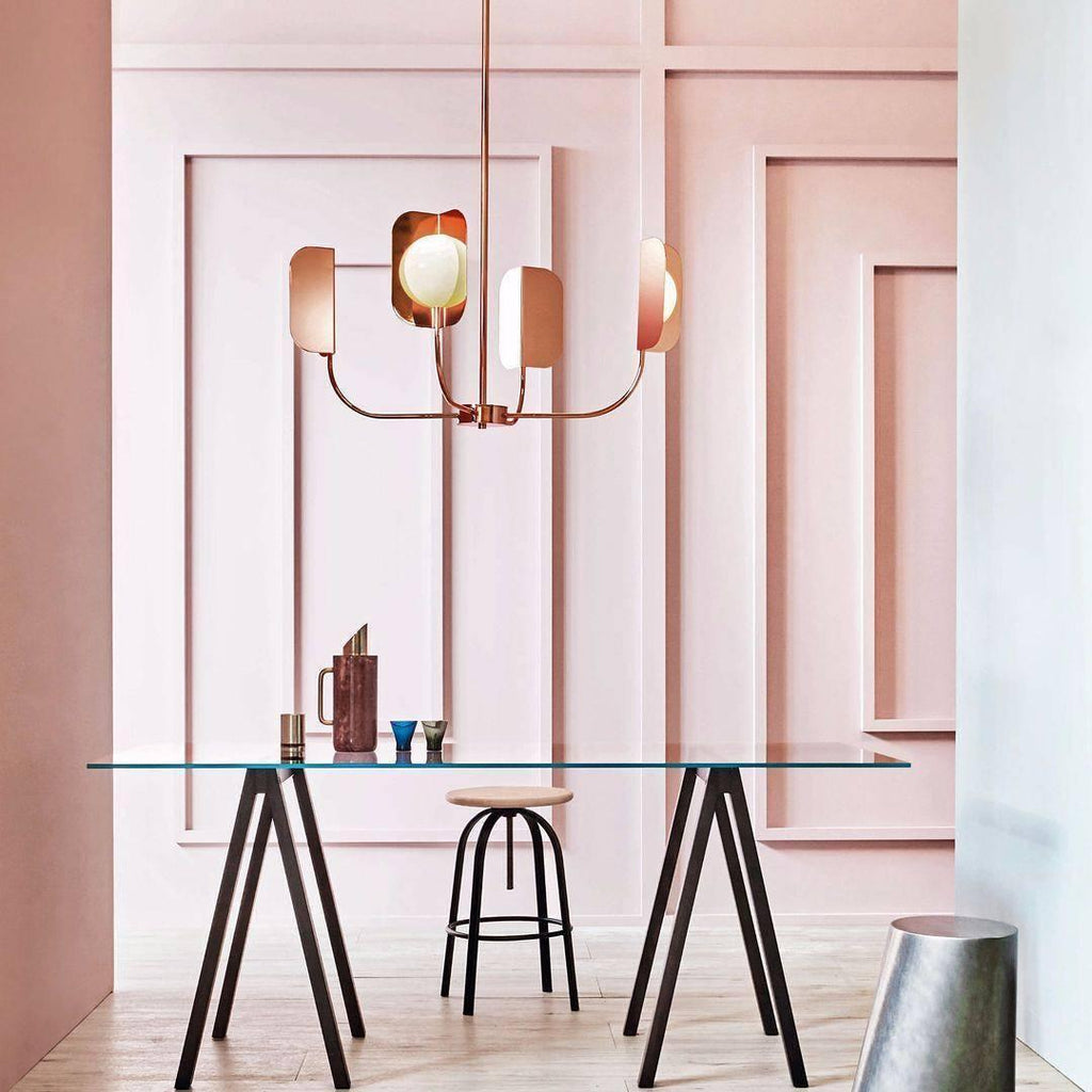 A small copper Clérachic chandelier with four leaf shades hangs in front of a pink panelled wall above a glass table and trestle legs.