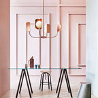 A small copper Clérachic chandelier with four leaf shades hangs in front of a pink panelled wall above a glass table and trestle legs.