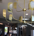 Looking up at a brass-toned Clérachic chandelier, with curved arms, leaf shades and a few bright globes against a recessed ceiling.