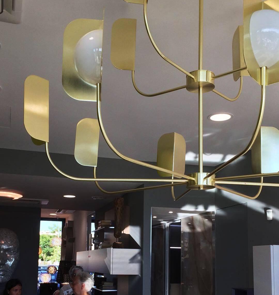 Looking up at a brass-toned Clérachic chandelier, with curved arms, leaf shades and a few bright globes against a recessed ceiling.