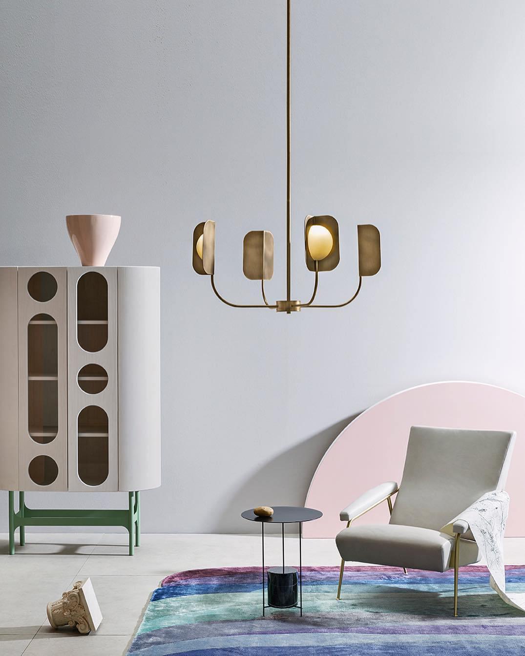 A small brass-toned Clérachic chandelier hangs in front of a tall cabinet with arched cut-outs, a pale armchair and a bright rug below.