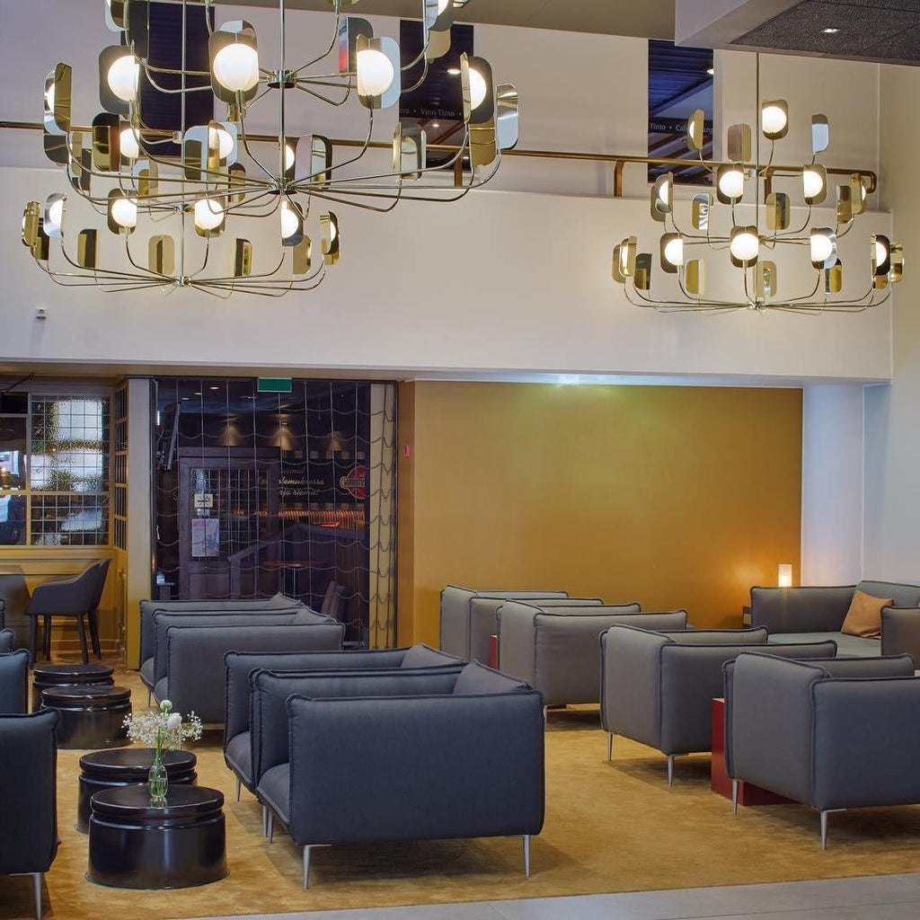 Two large brass-toned Clérachic chandeliers lit in a hotel lounge, with grey armchairs, low round tables and a gold-toned wall.
