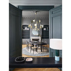 A brass-toned Clérachic chandelier hangs in a dark green panelled room, seen through double doors above an oval table with black chairs.
