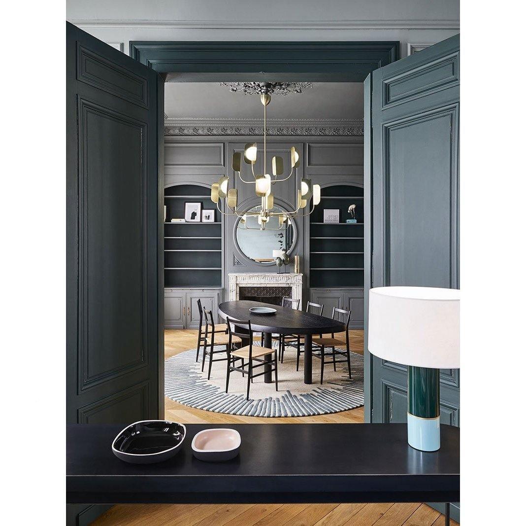 A brass-toned Clérachic chandelier hangs in a dark green panelled room, seen through double doors above an oval table with black chairs.