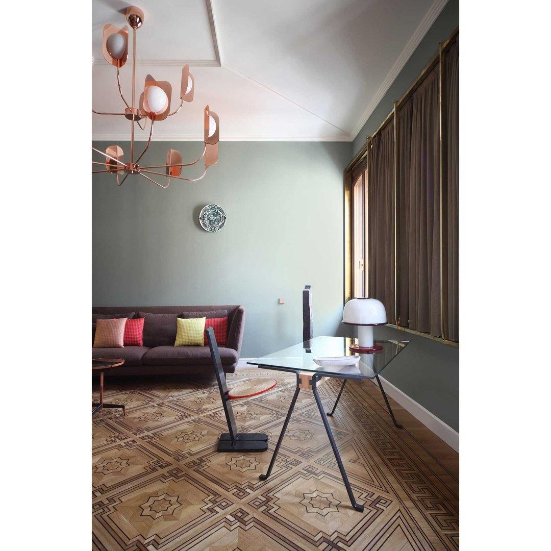 A copper Clérachic chandelier in a grey-green room with a patterned floor, a glass desk and a red sofa under a window.