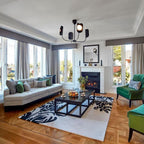 A black Clérachic chandelier with five leaf shades hangs in a bright sitting room with a fireplace, green armchairs and a patterned rug.