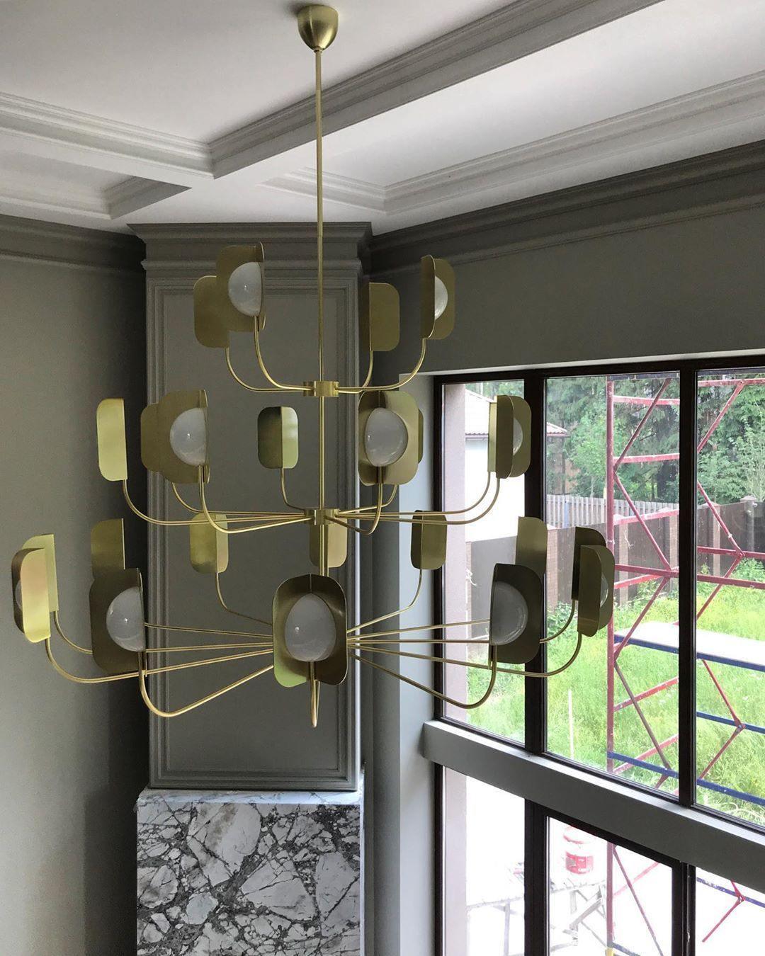 A tall brass-toned Clérachic chandelier with many lit leaf shades hangs in a stairwell with black window frames and a marble plinth.