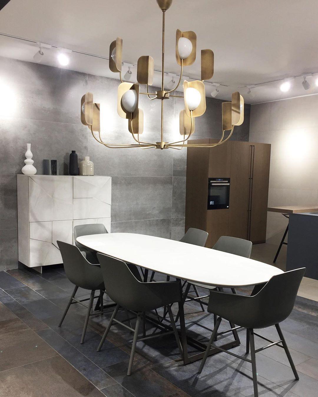 A brass-toned Clérachic chandelier hangs over an oval white table and grey chairs in a kitchen-diner with a stone-clad wall.