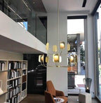 A brass-toned Clérachic chandelier in a double-height room with open shelves, glass balustrades and a rust-coloured armchair.