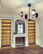 A Clérachic chandelier with five dark leaf shades hangs from a ceiling rose in front of a fireplace flanked by lit wooden shelves.