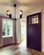 A Clérachic chandelier hangs in a panelled room with herringbone floors and a dark door, its black leaf shades lit softly from within.