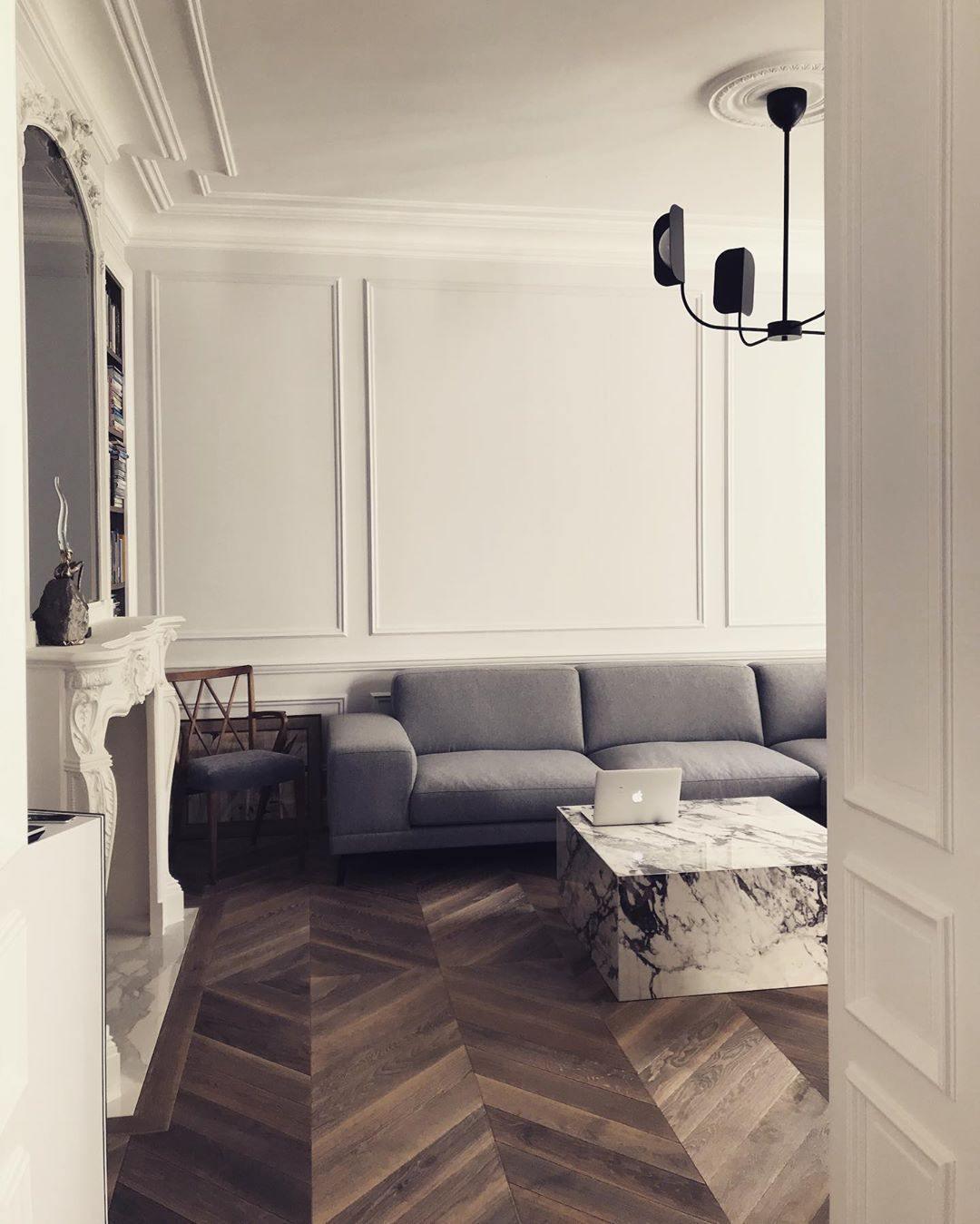 A small black Clérachic chandelier hangs in a hall with white wall panelling and a marble coffee table, beside a grey sofa.