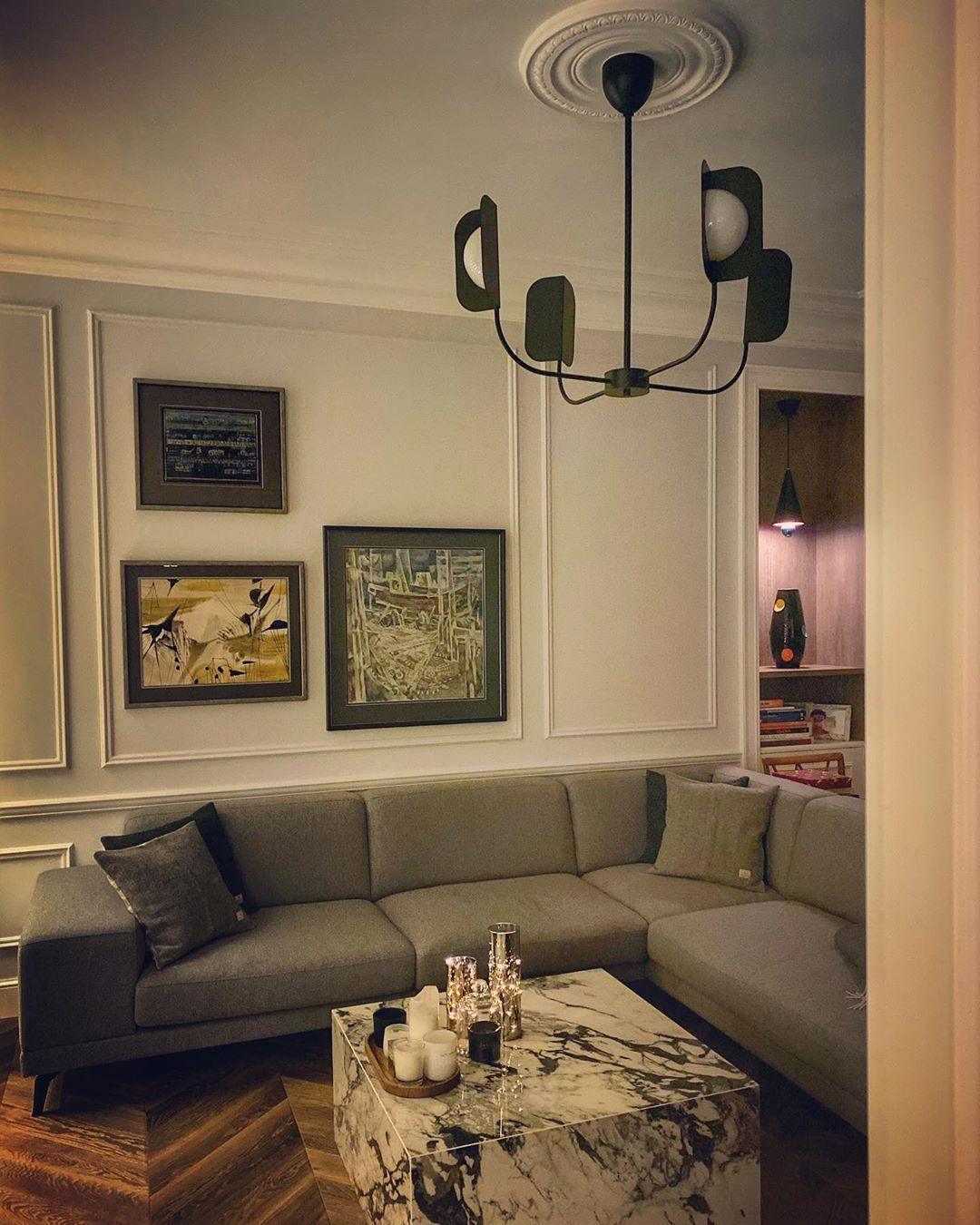 A black Clérachic chandelier with four leaf shades hangs above a grey sofa and marble table in a room with framed art.