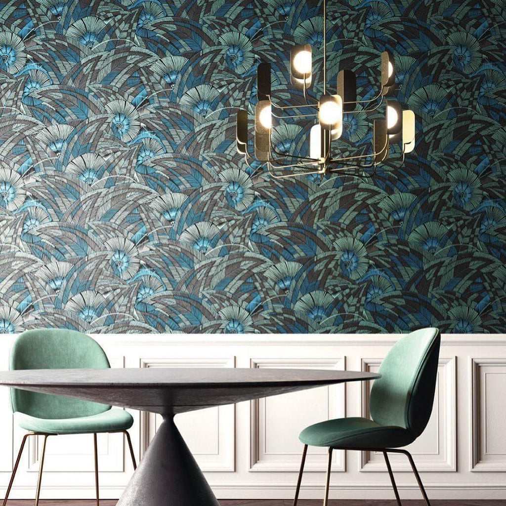 A brass-toned Clérachic chandelier lit beside a wall of tropical blue-green wallpaper, above a dark oval table with green chairs.