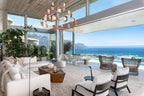 A copper Clérachic chandelier with lit leaf shades hangs under a timber ceiling in a seaside living room with glass walls and ocean views.