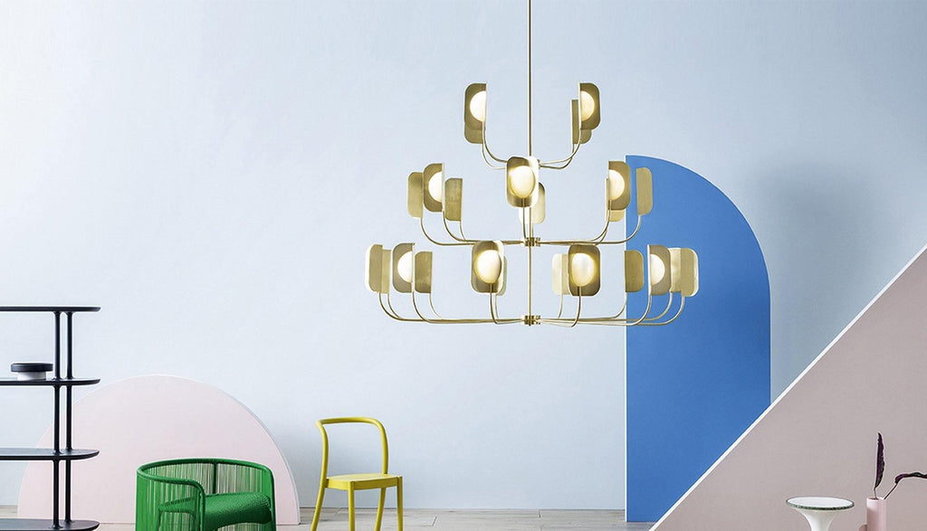 A brass-toned Clérachic chandelier of three tiers hangs against a pale blue wall with an arched blue panel and bright chairs below.