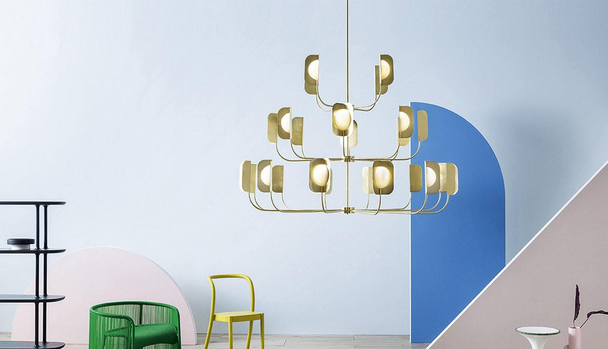 A brass-toned Clérachic chandelier of three tiers hangs against a pale blue wall with an arched blue panel and bright chairs below.