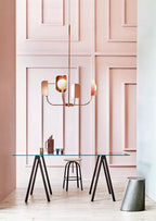 A small copper Clérachic chandelier hangs in front of a tall pink panelled wall, above a glass table on trestle legs and a stool.