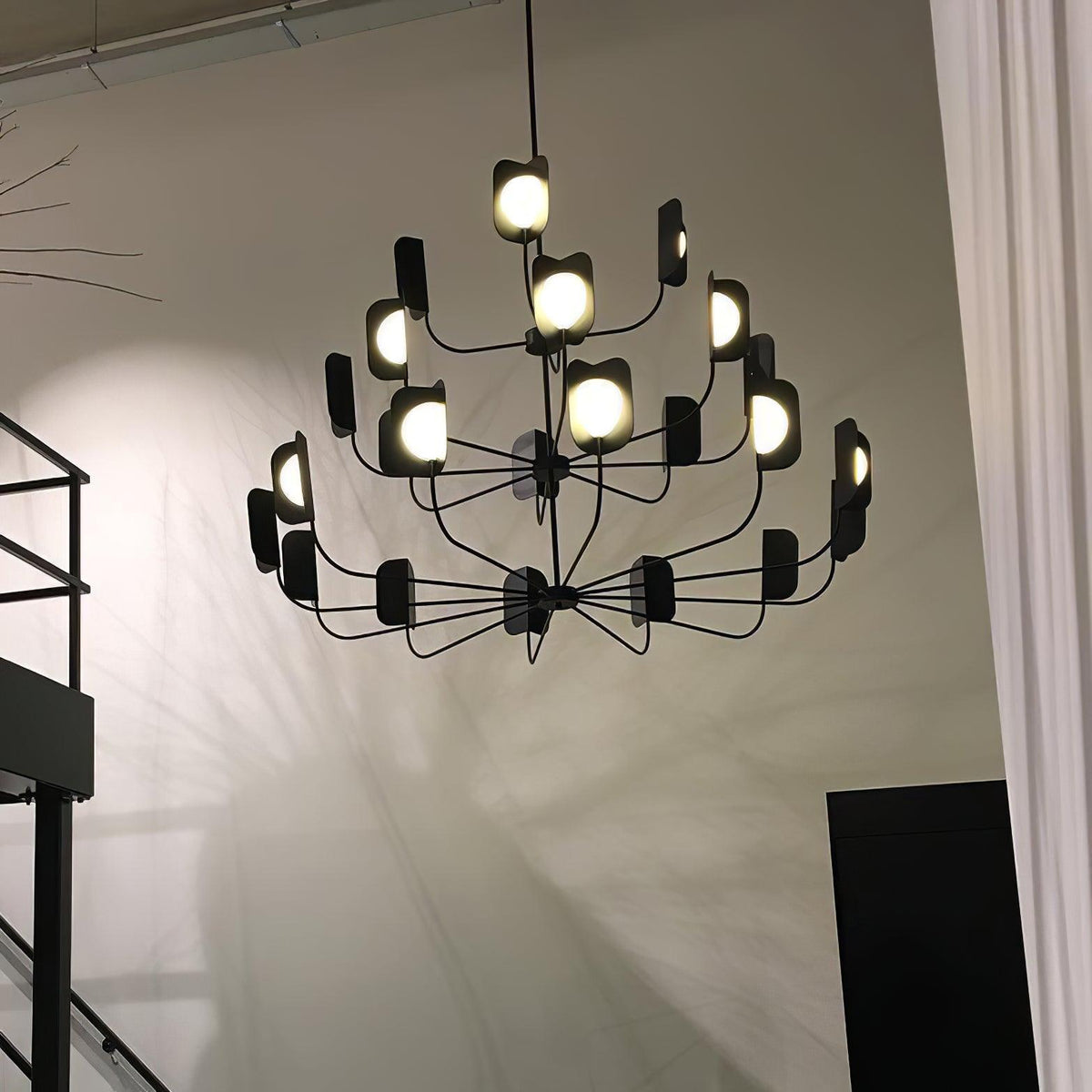 A black Clérachic chandelier of three tiers glowing in a double-height space beside a black steel stair and a pale wall.