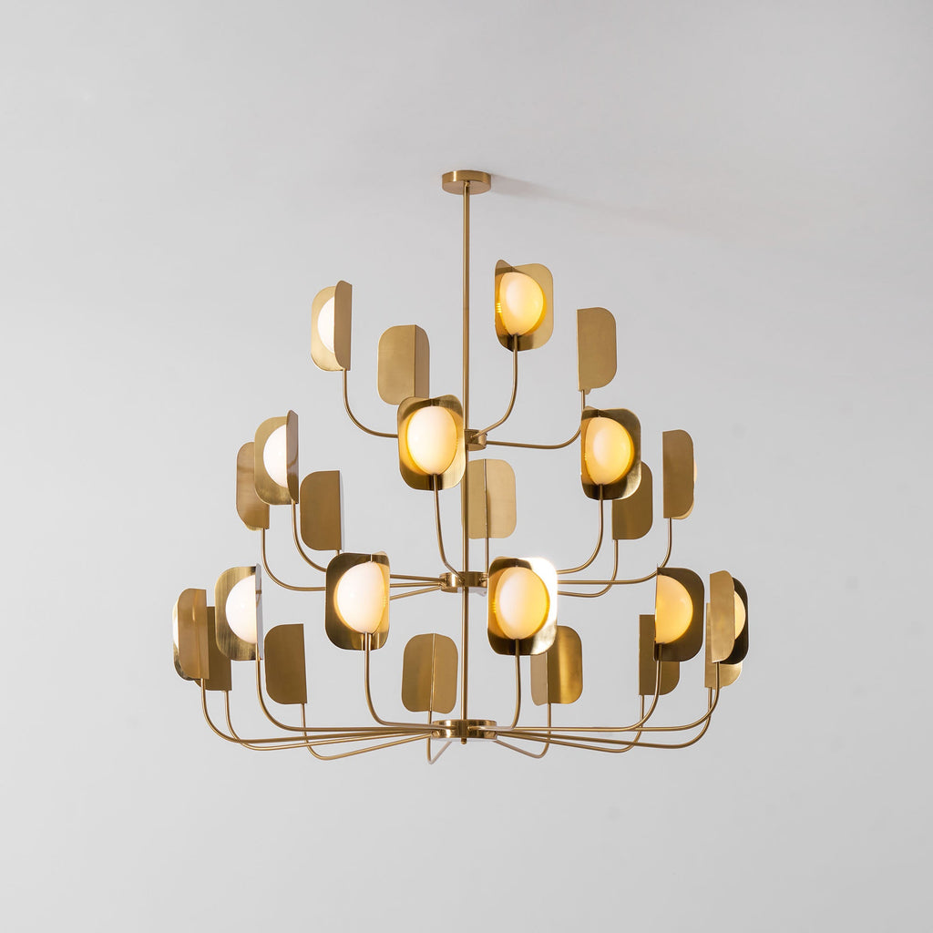 A brass-toned Clérachic chandelier of three tiers with lit leaf shades and globes, on a pale grey backdrop.