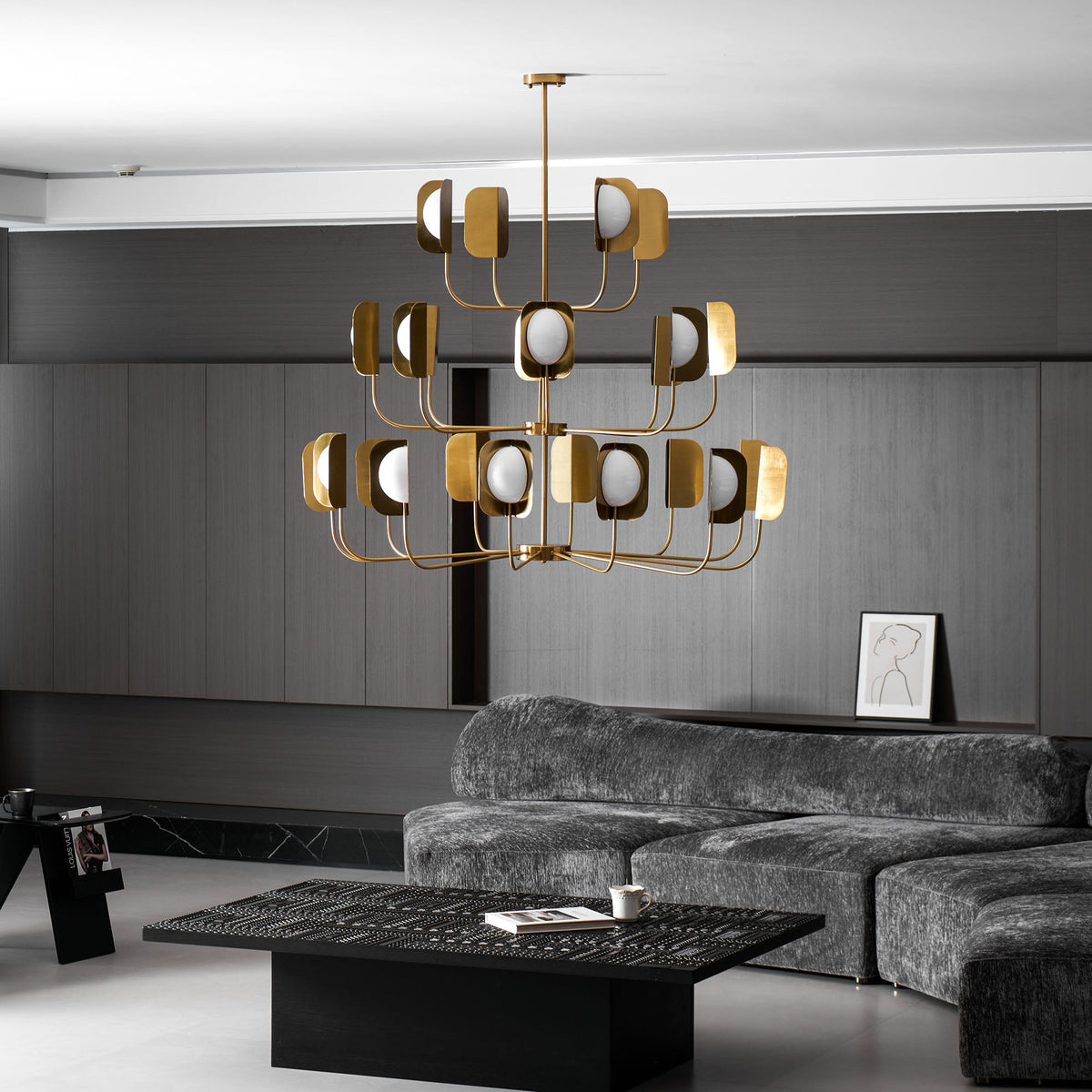 A large brass-toned Clérachic chandelier hangs above a low black coffee table in a dark panelled lounge with a grey sofa.