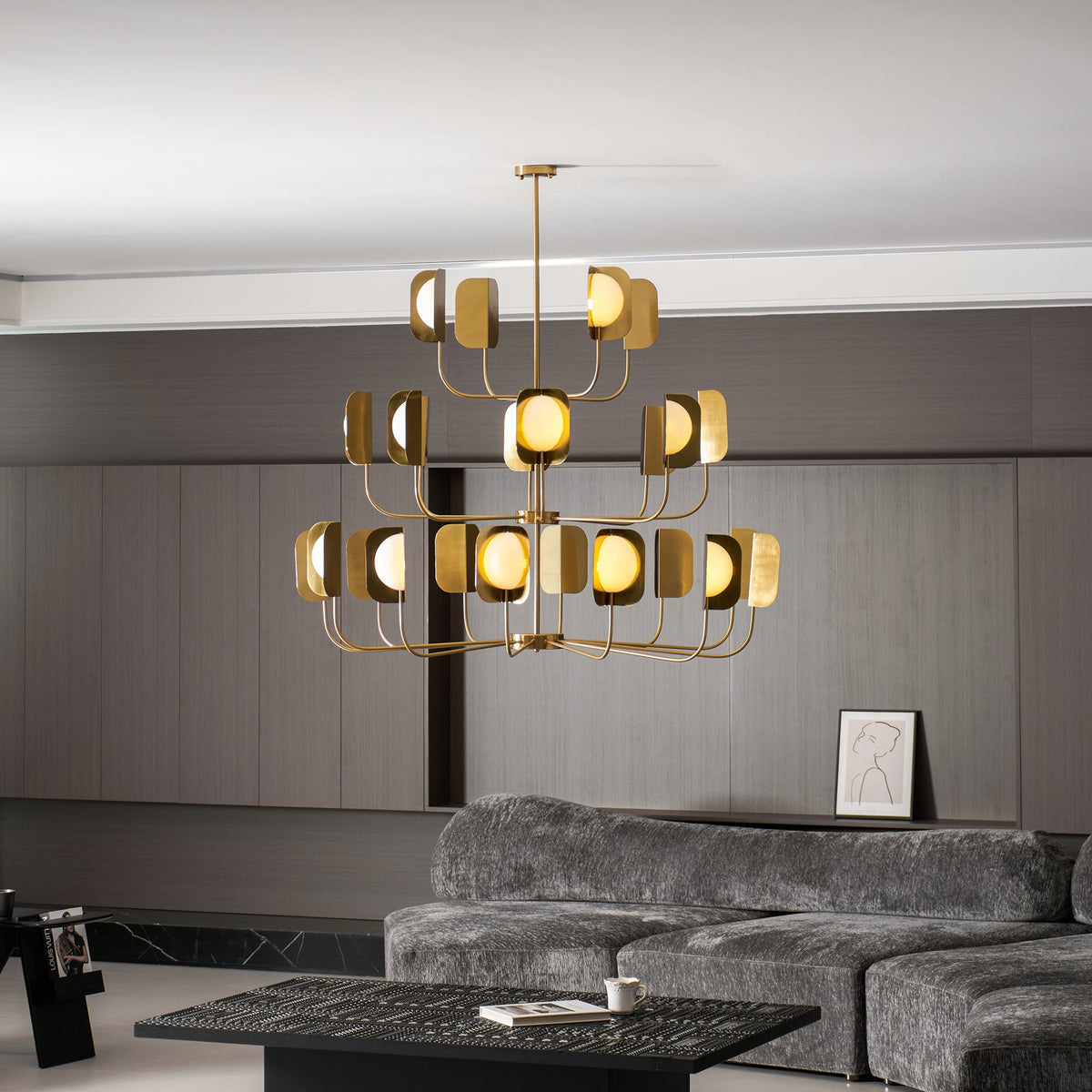 A large brass-toned Clérachic chandelier with three tiers of arms, hung in a grey lounge above a low black table and a sofa.